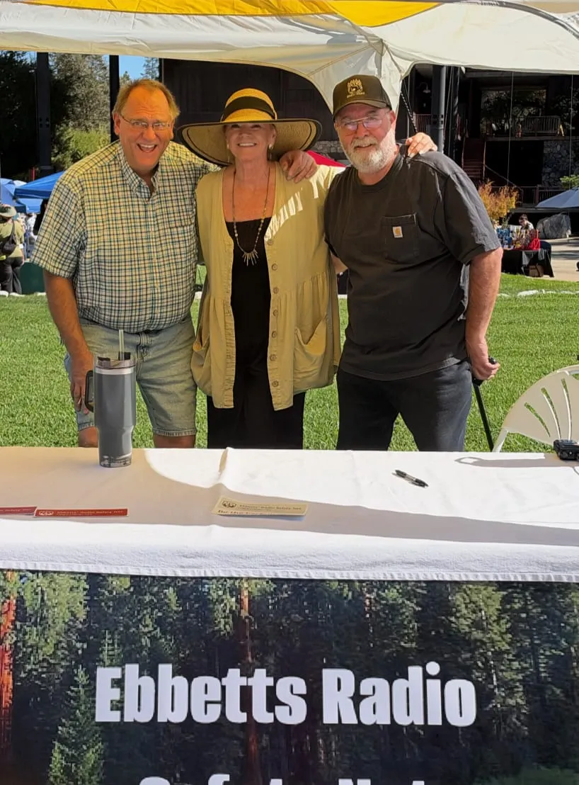 Kenny, Marlaine, and Mike smiling arm in arm behind the Ebbetts Radio Safety Net table on a sunny day